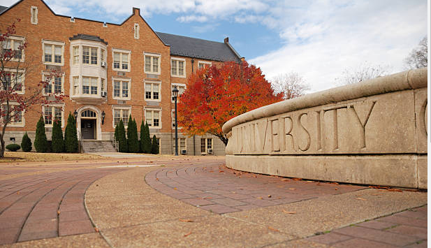 A higher education campus shows a low stone wall in the foreground engraved with the name of the university leading to a n academic building in the background.