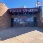Pueblo ice arena glass door entrance in daytime. The entrance is recessed into a stone wall on one side and a stone arch on the other side. Sign at entrance reads 'Pueblo Ice Arena: Welcome to!'