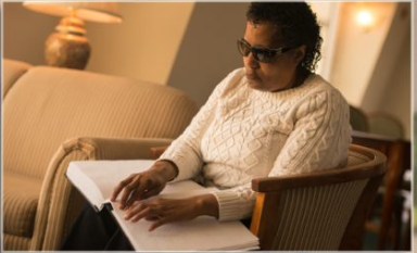A visually impaired woman wearing dark glasses sits in a sofa and runs her fingers over the pages in a Braille book. This is one of the accessibility features provided at the Georgia Library Service for the Blind and Print-disabled.